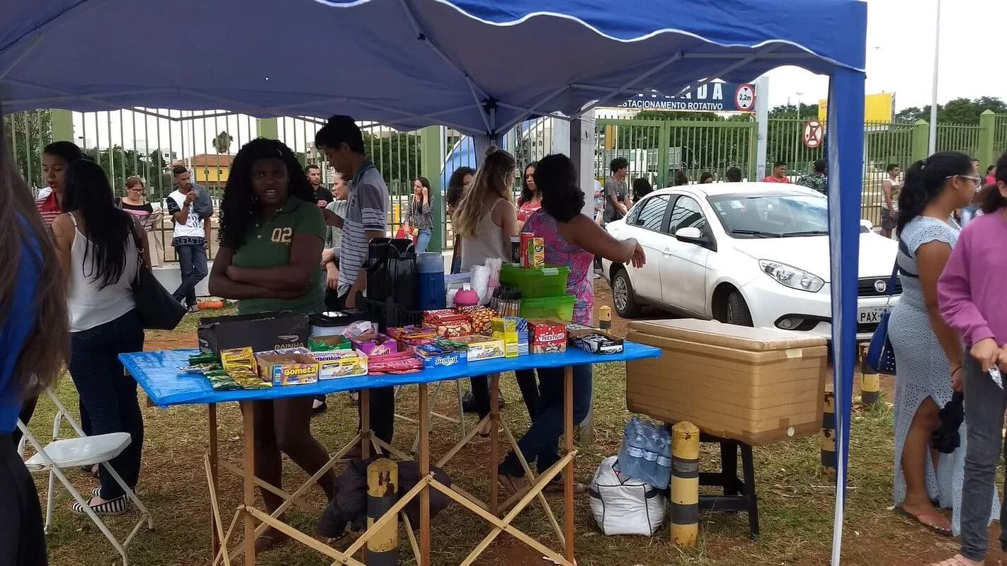Candidatos reunidos em área externa próxima a local de prova, com barracas de vendedores ao redor