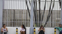 Candidatos sentados em banco de concreto aguardam do lado de fora de local de prova