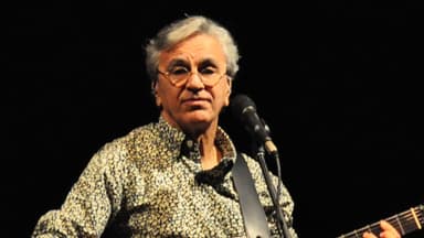 Caetano Veloso tocando violão durante show, com microfone à frente