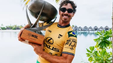 Yago Dora sorri segurando o troféu de campeão mundial de surfe de 2025, vestindo camisa amarela da seleção brasileira, com bangalôs sobre a água ao fundo em Fiji