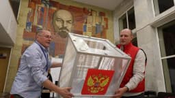 Dois funcionários carregam uma urna eleitoral transparente com o brasão da Rússia em seção de votação.