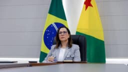 Mailza Assis em ambiente institucional, sentada entre as bandeiras do Brasil e do Acre