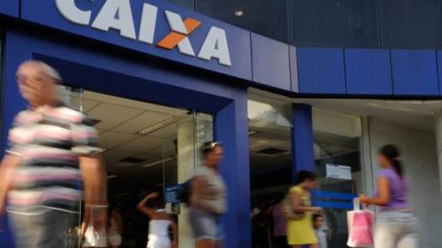 Fachada de agência bancária da Caixa com pessoas passando na calçada