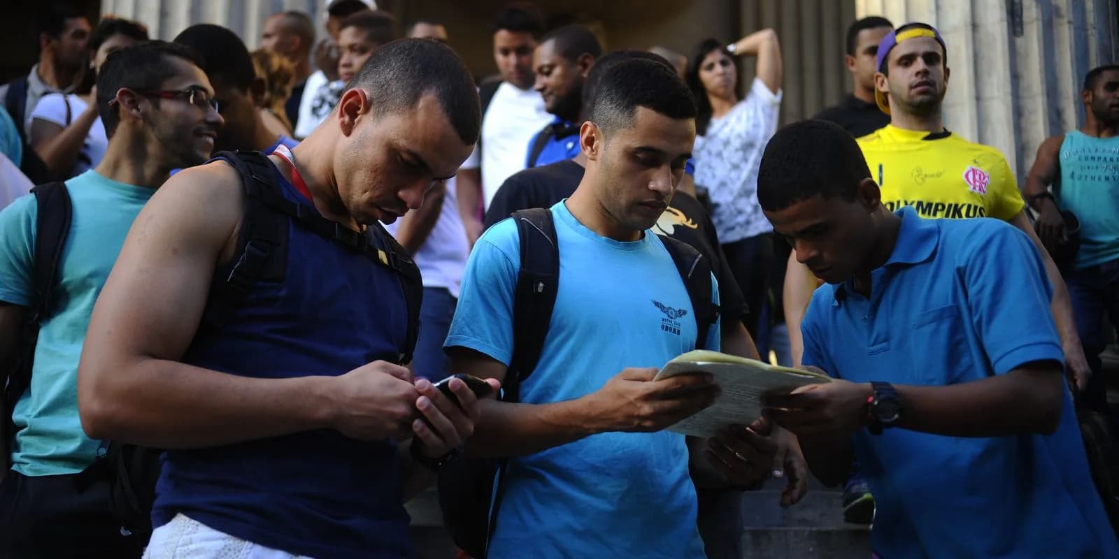 Grupo de candidatos consultando documentos e celular em fila do lado de fora de prédio público, foto de arquivo ilustrativa de concurso público