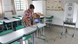 Mulher prepara cabine de votação em sala de aula com urna eletrônica, sob placa da Justiça Eleitoral
