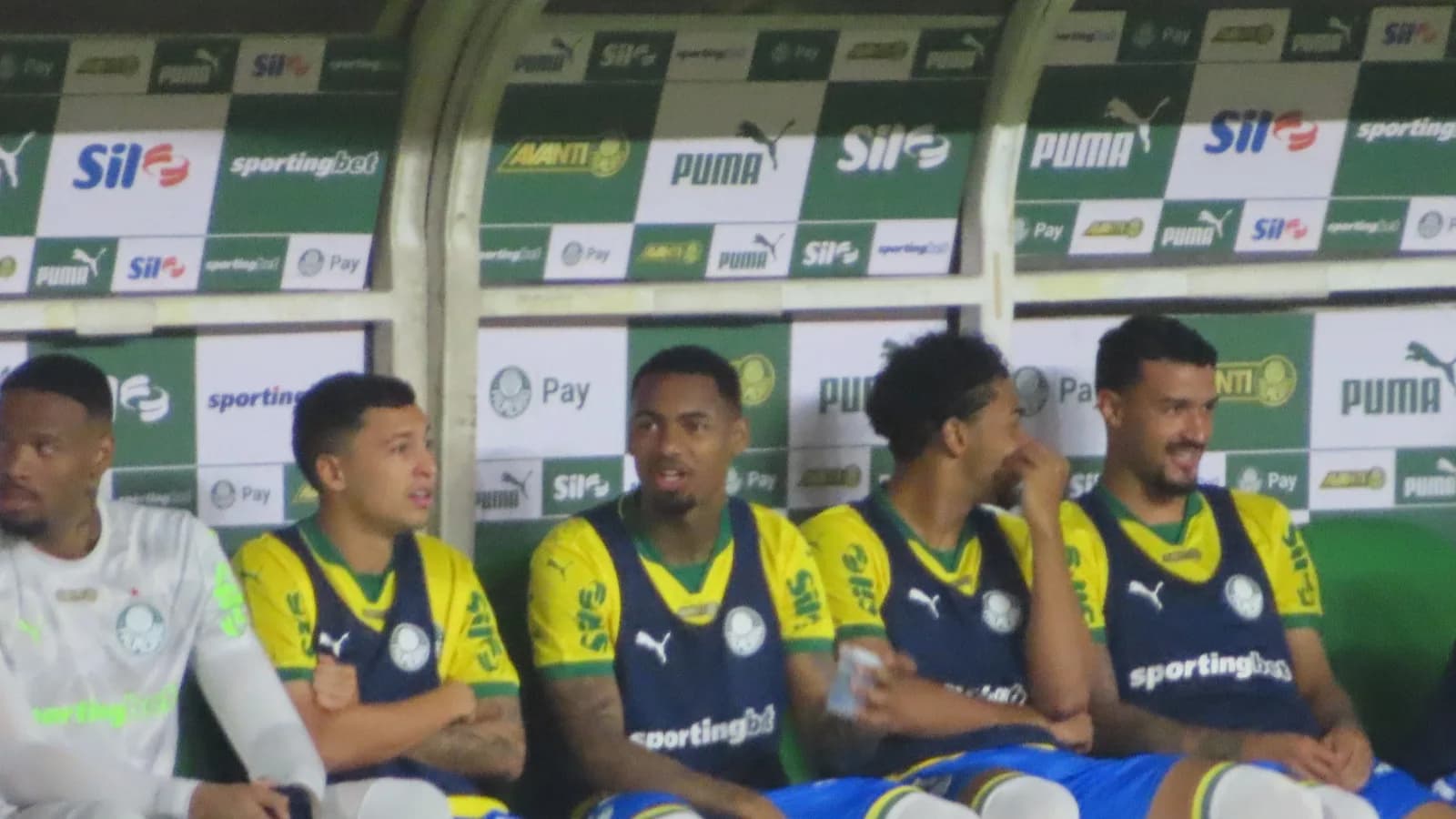 Jefté sentado no banco de reservas do Palmeiras ao lado de outros jogadores, com uniforme reserva amarelo e azul