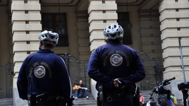 Dois guardas civis metropolitanos de costas, com capacete de ciclista, em patrulha em frente a prédio histórico