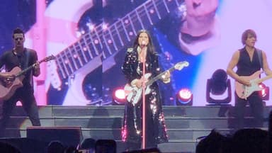 Laura Pausini se apresenta ao microfone durante show, ladeada por guitarristas