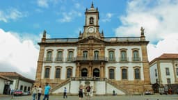 Fachada do Museu da Inconfidência, com torre e relógio, em Ouro Preto, com visitantes caminhando em frente ao prédio. Imagem de arquivo.