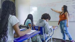 Candidatos em sala de aula durante prova, com professora escrevendo no quadro branco