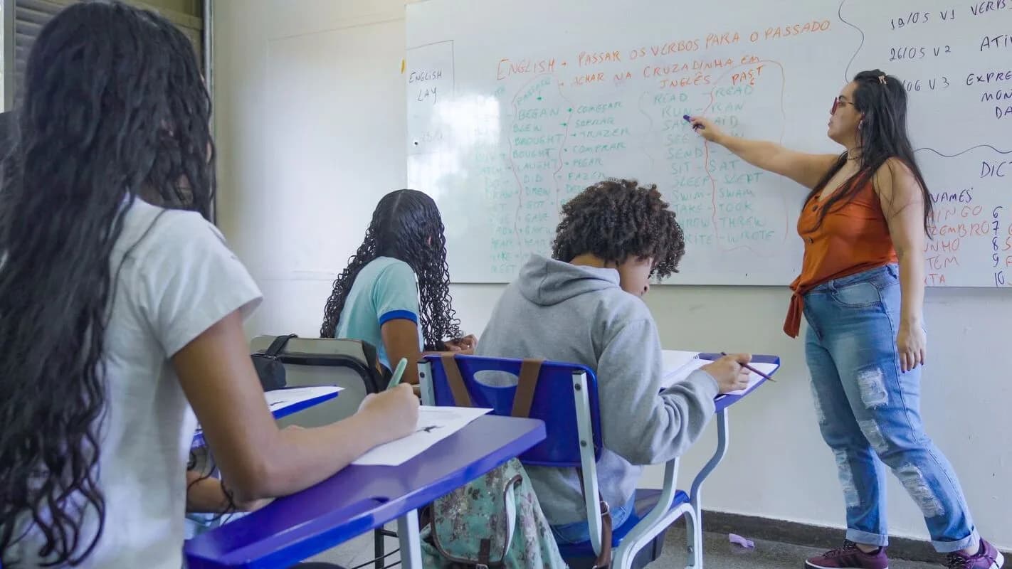 Candidatos em sala de aula durante prova, com professora escrevendo no quadro branco