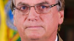 Jair Bolsonaro em retrato sério, de óculos e terno, com a bandeira do Brasil ao fundo