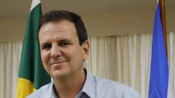 Eduardo Paes sorri em retrato oficial, com bandeiras do Brasil e do Rio de Janeiro ao fundo