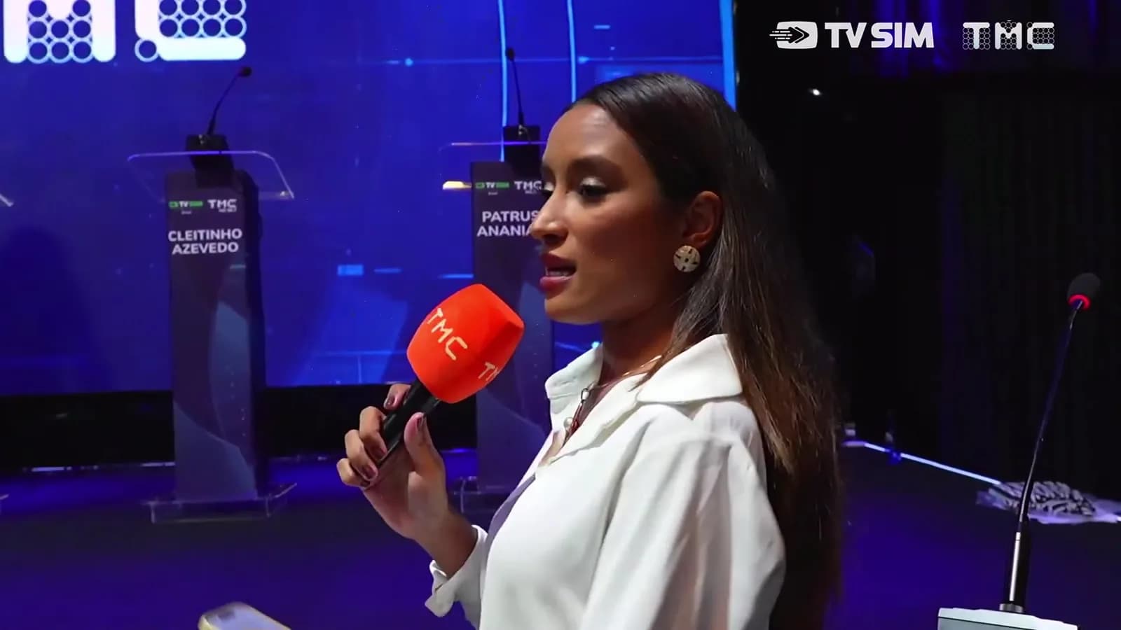 Repórter com microfone da TMC em pé diante de púlpitos vazios no palco do debate, identificados com os nomes 'Cleitinho Azevedo' e 'Patrus Ananias'