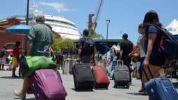 Imagem de arquivo de turistas caminhando com malas de rodinha em área portuária, com um navio de cruzeiro atracado ao fundo