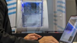 Pessoa trabalha em laptop ao lado de computador com componentes expostos, em evento de tecnologia