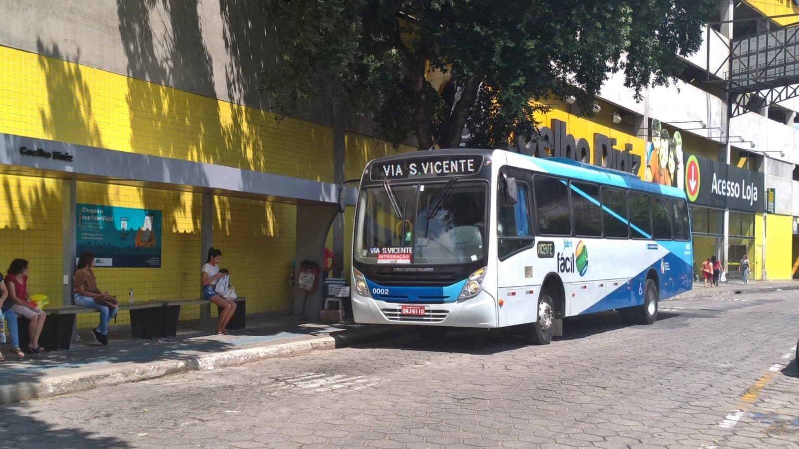 Ônibus da Saritur parado em ponto de transporte coletivo, com passageiras aguardando no banco ao lado