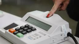 Mão com unha vermelha pressiona botão de urna eletrônica durante votação