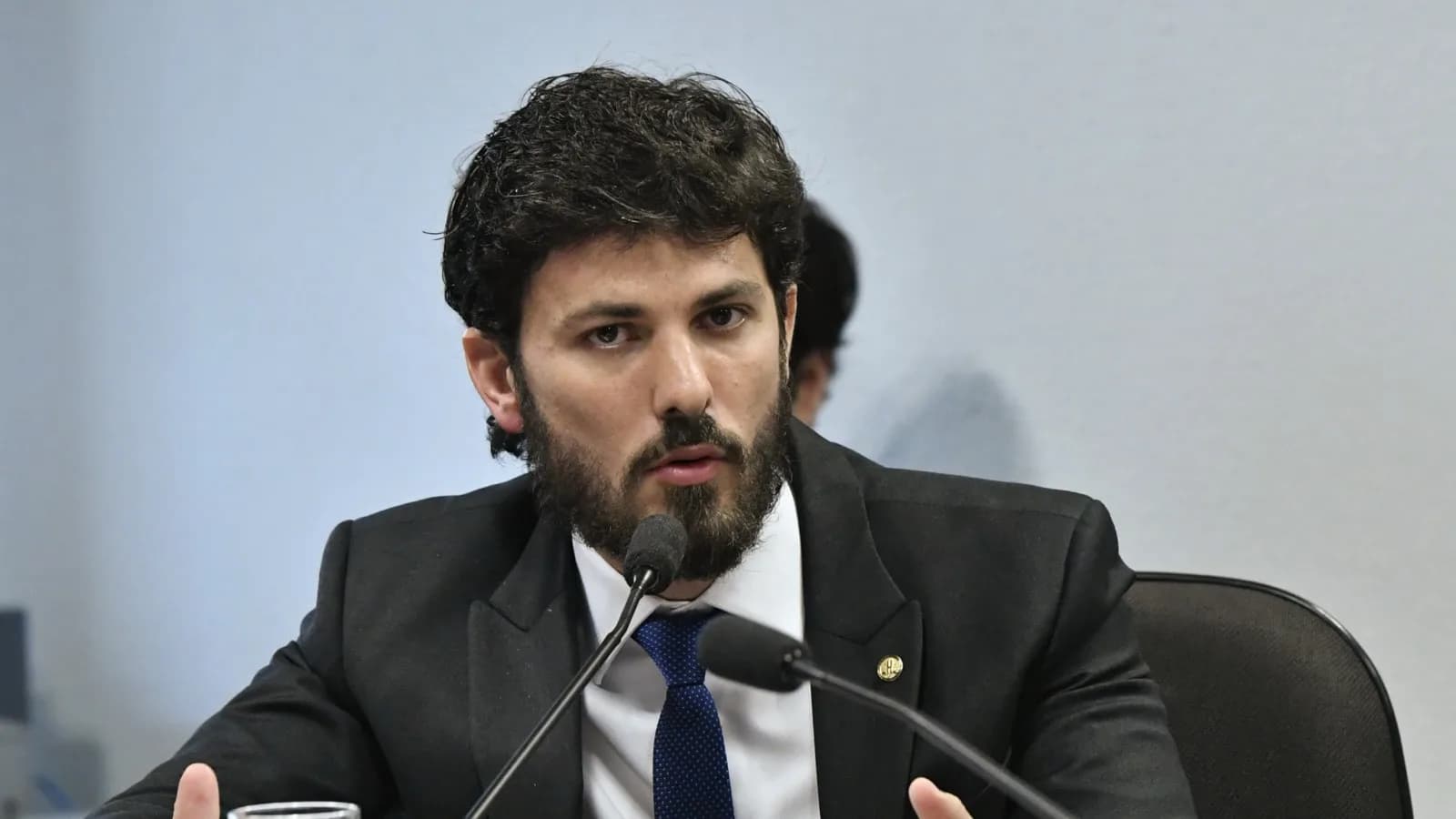Marcelo Aro fala ao microfone em audiência pública, de terno escuro e barba