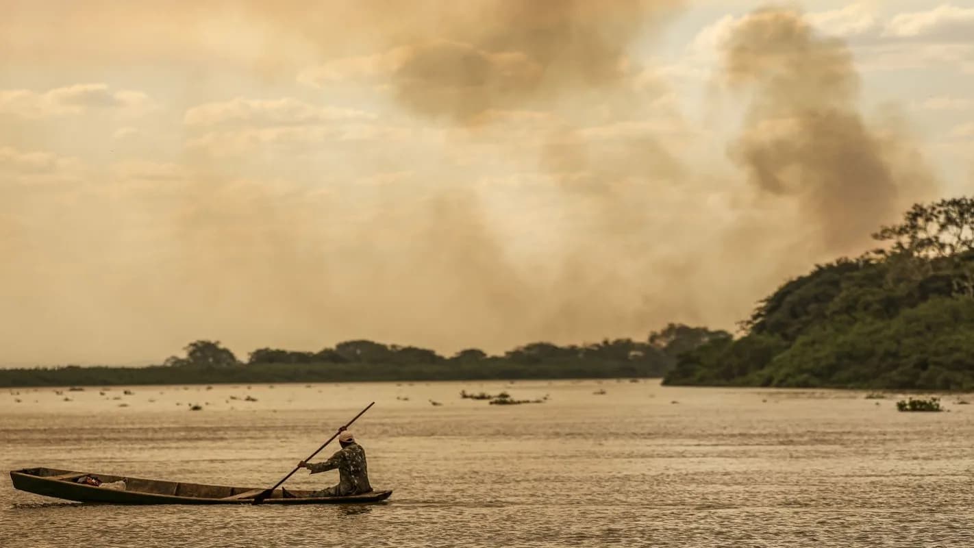 Homem rema em canoa em rio da Amazônia com fumaça de queimada ao fundo