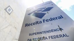 Placa de sinalização com o logotipo da Receita Federal e os dizeres Ministério da Fazenda e Superintendência da Receita Federal, fotografada de baixo para cima contra o céu, em Brasília.