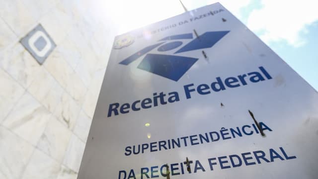 Placa de sinalização com o logotipo da Receita Federal e os dizeres Ministério da Fazenda e Superintendência da Receita Federal, fotografada de baixo para cima contra o céu, em Brasília.