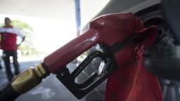 Foto de arquivo: bico de bomba de combustível abastecendo um veículo em um posto, com frentista de uniforme vermelho desfocado ao fundo