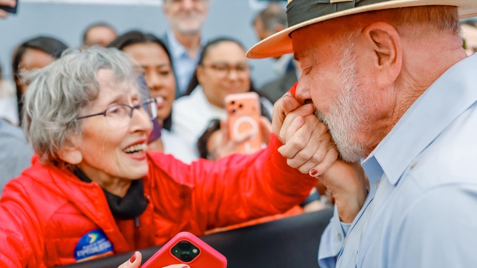 O presidente Lula, de chapéu de palha, beija a mão de uma apoiadora idosa de casaco vermelho, cercado por outras pessoas, durante visita a Belo Horizonte em junho de 2026