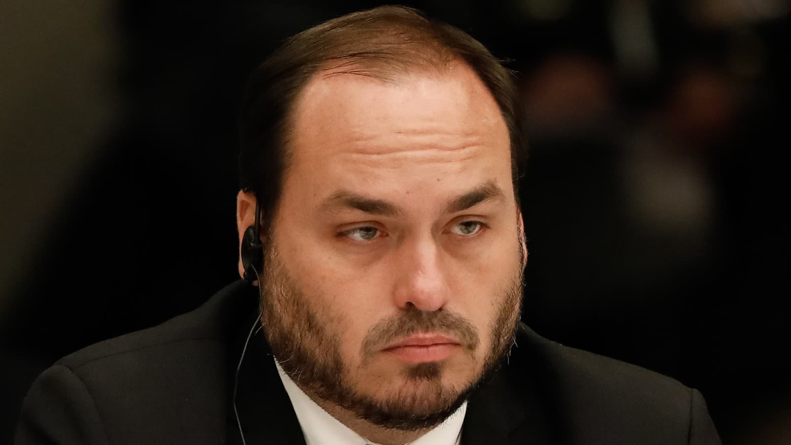 Carlos Bolsonaro sentado à mesa de reunião, de terno escuro e gravata, com ponto de tradução simultânea na orelha; imagem de arquivo de 2022