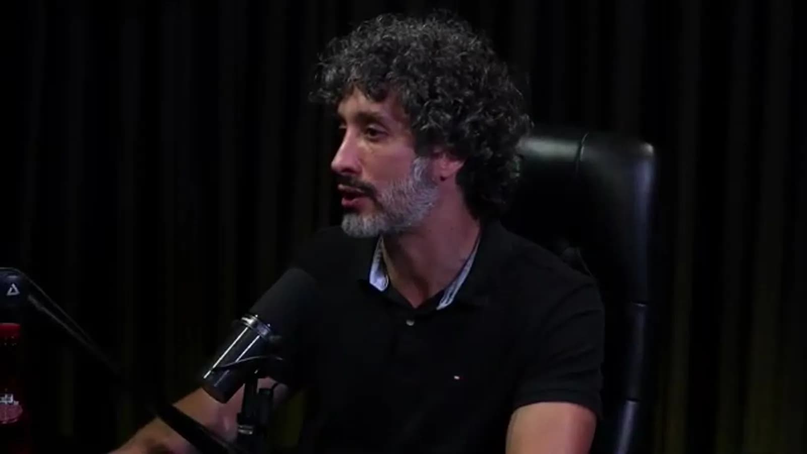 Carlos Eduardo Grimaldi fala recostado na cadeira, em perfil levemente lateral, em estúdio de podcast com cortina escura ao fundo