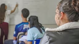Professora escreve no quadro enquanto estudantes aparecem de costas em sala de aula; foto de arquivo, não da convocação em Uberaba.