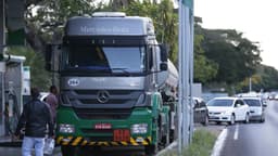 Caminhão-tanque parado em posto de combustíveis à beira de uma via, com pessoas ao lado e fila de carros ao fundo. Imagem de arquivo.