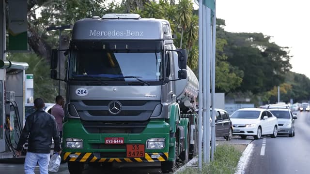 Caminhão-tanque parado em posto de combustíveis à beira de uma via, com pessoas ao lado e fila de carros ao fundo. Imagem de arquivo.