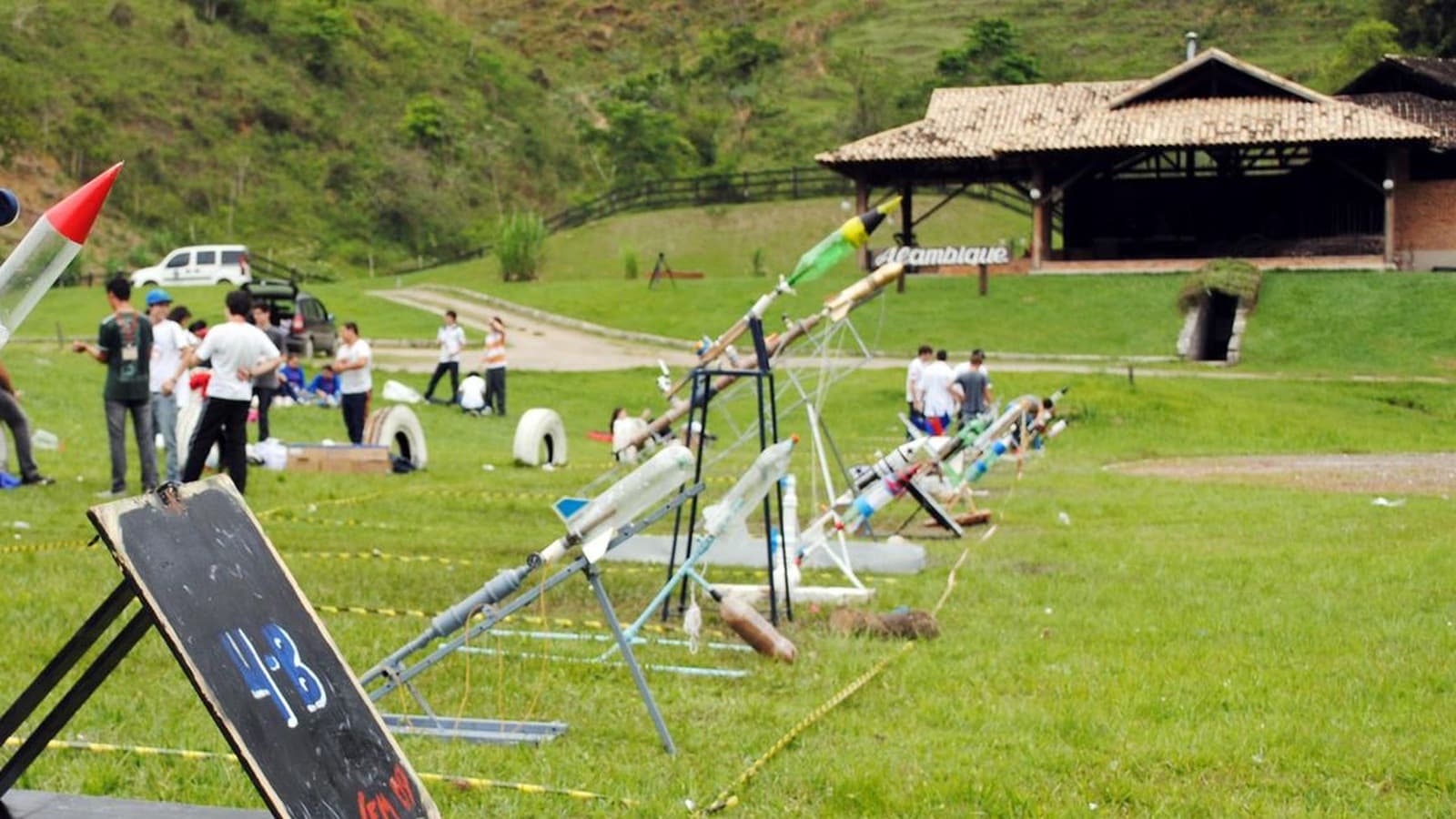 Foguetes coloridos alinhados em rampas de lançamento em um campo, com estudantes ao fundo
