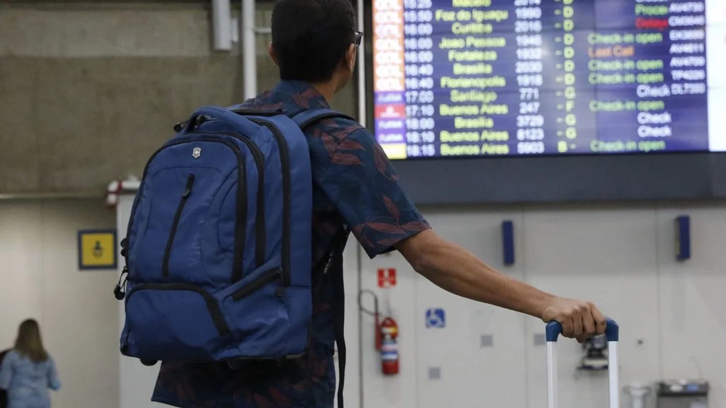 Viajante de costas com mochila e mala observa painel de voos em aeroporto