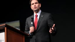 Marco Rubio discursando ao microfone em um púlpito, de terno escuro e gravata vermelha