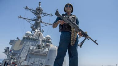 Militar da Marinha dos EUA em pé no convés de um contratorpedeiro no Golfo de Omã, empunhando uma metralhadora, com o navio ao fundo