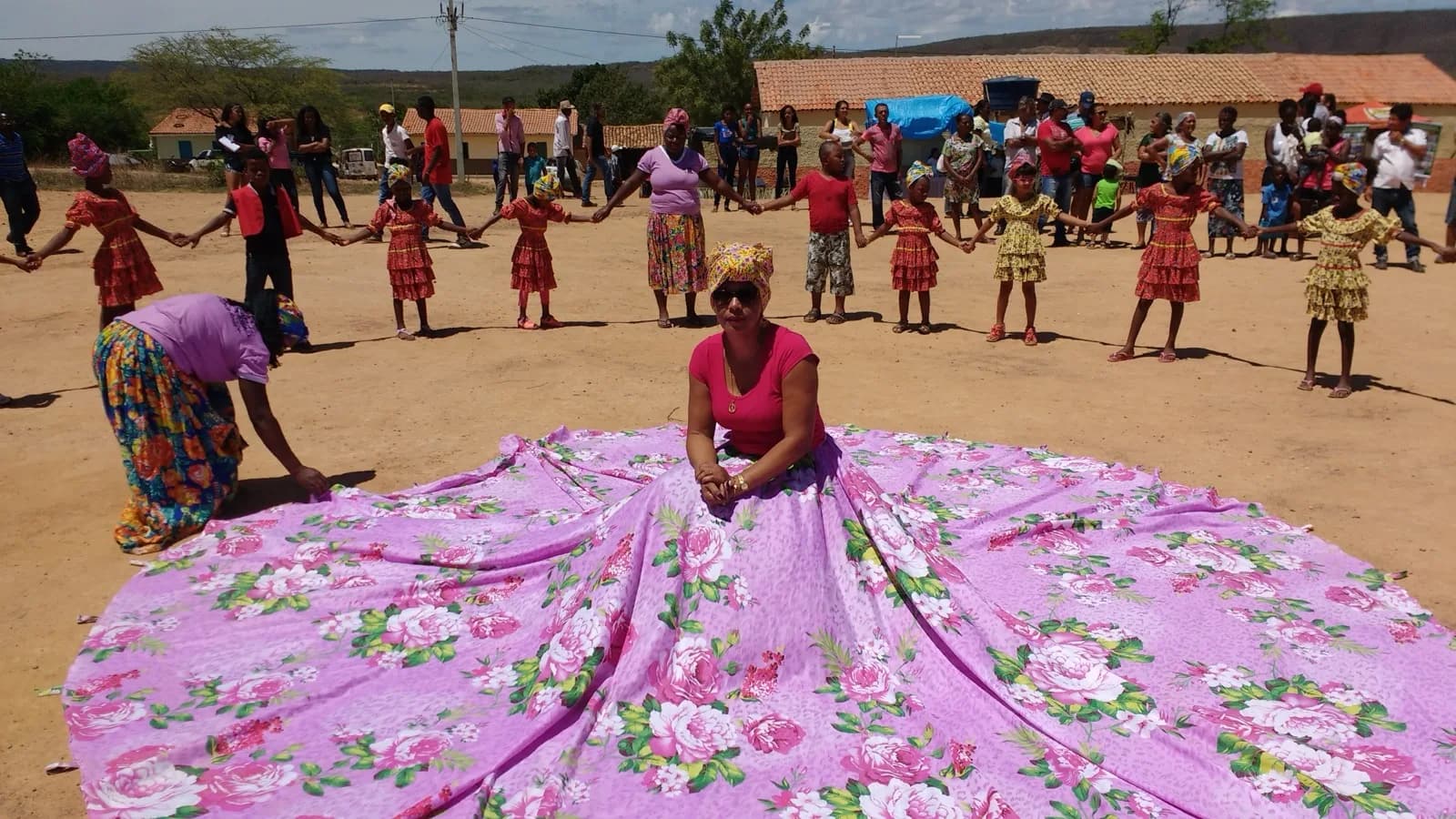 Mulheres e crianças quilombolas em roda durante encontro comunitário, com mulher em vestido tradicional ao centro