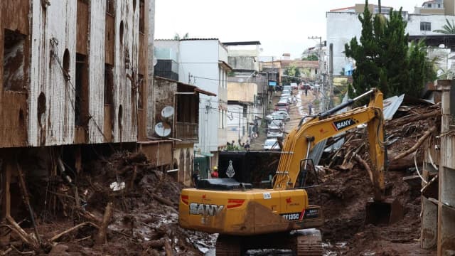 Rua destruída após as enchentes de fevereiro de 2026 em Minas Gerais, com escavadeira removendo escombros