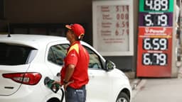 Frentista de uniforme vermelho abastece um carro branco em posto de combustível, com o painel de preços ao fundo; imagem de arquivo