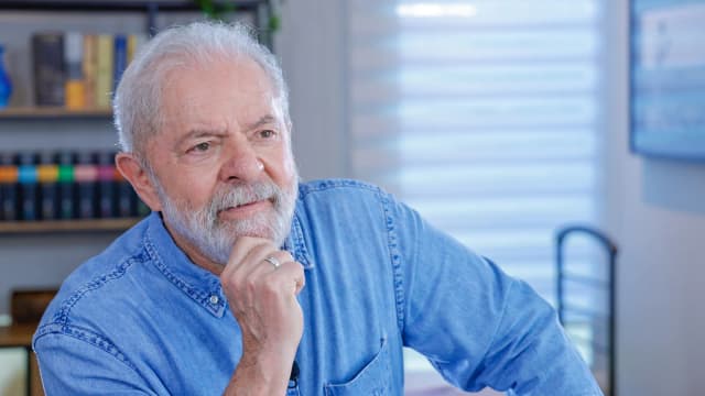 Lula gesticula durante entrevista, com a mão no queixo, vestindo camisa jeans azul