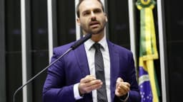 Eduardo Bolsonaro discursa na tribuna da Câmara dos Deputados, de terno azul, diante de bandeira do Brasil