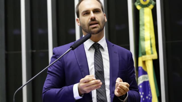 Eduardo Bolsonaro discursa na tribuna da Câmara dos Deputados, de terno azul, diante de bandeira do Brasil