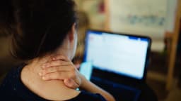 Mulher segura o pescoço com dor enquanto olha para a tela do notebook