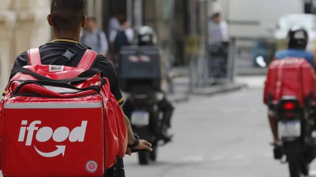 Entregador de aplicativo com bolsa térmica nas costas ao lado de motocicletas em rua movimentada