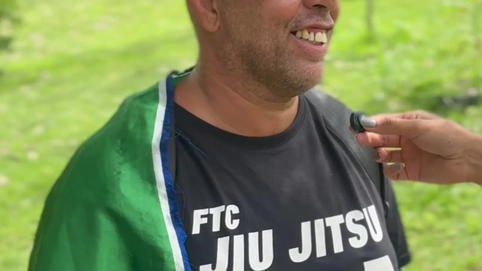 Marcos de Oliveira, professor de jiu-jítsu, dá entrevista à TV Sim Brasil usando camisa do FTC Jiu Jitsu USA com adesivo da campanha de Flávio Bolsonaro e bandeira verde sobre os ombros
