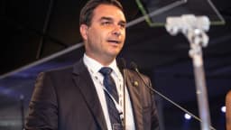 Flávio Bolsonaro discursa em pódio durante evento, vestindo terno escuro e crachá de credenciamento