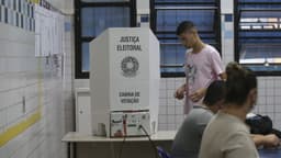 Cabina de votação da Justiça Eleitoral com urna eletrônica sobre a mesa, eleitor se aproximando e mesários sentados em uma seção eleitoral. Imagem de arquivo