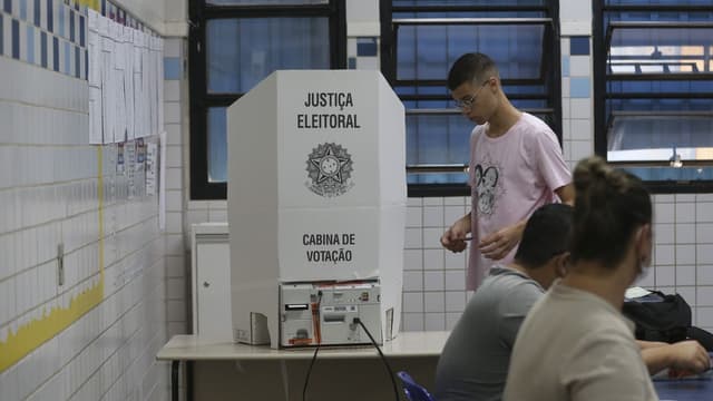 Cabina de votação da Justiça Eleitoral com urna eletrônica sobre a mesa, eleitor se aproximando e mesários sentados em uma seção eleitoral. Imagem de arquivo