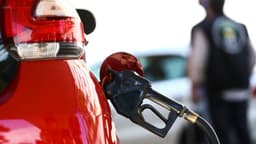 Bico de bomba de combustível abastecendo um carro vermelho em posto de Brasília, com fiscal desfocado ao fundo; imagem de arquivo